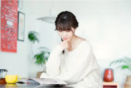 テーブルに座って複数の資料を比較検討している白いセーターの女性。コーヒーカップとスマートフォンが置かれ、集中して講座選びをしている様子