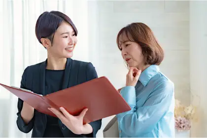 黒いスーツを着た女性と水色のシャツを着た女性が赤いファイルを一緒に見ながら笑顔で相談している様子の写真
