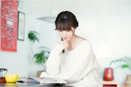 テーブルに座って複数の資料を比較検討している白いセーターの女性。コーヒーカップとスマートフォンが置かれ、集中して講座選びをしている様子