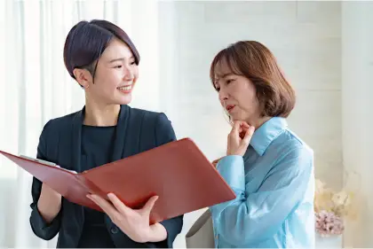 黒いスーツを着た女性と水色のシャツを着た女性が赤いファイルを一緒に見ながら笑顔で相談している様子の写真