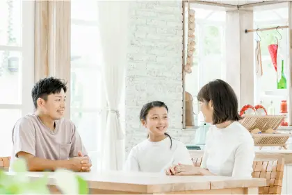 明るいリビングで父親、母親、子供の3人家族がテーブルを囲んで資料を見ながら和やかに話し合っている様子の写真